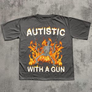 May include: Dark gray t-shirt with the text "AUTISTIC" above a skeleton figure holding two guns, surrounded by flames, and the text "WITH A GUN" below.