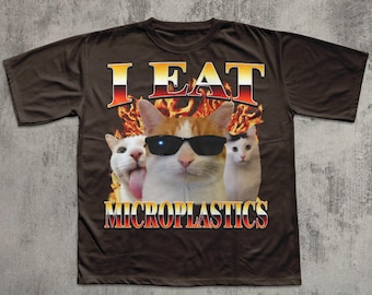 May include: Brown t-shirt with a graphic featuring three cats. The central cat wears sunglasses. Text above reads "I EAT" in a fiery font, and below, "MICROPLASTICS" in a similar style. The design includes flames.