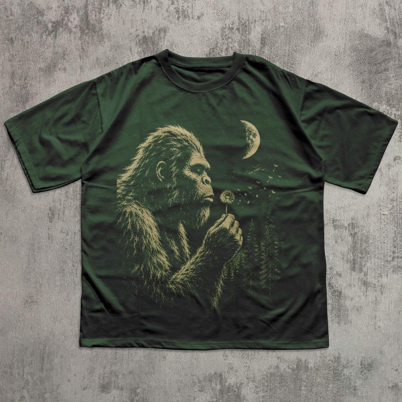 May include: A dark green short-sleeve t-shirt featuring a graphic print of a hairy creature resembling a Bigfoot blowing on a dandelion. The design includes a crescent moon and a forest silhouette in the background, rendered in muted light green tones.