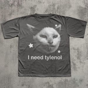 May include: Dark gray t-shirt featuring a white, pixelated cat face with three white stars. The text "I need tylenol" is printed in white below the cat graphic. The shirt is made of a soft material.