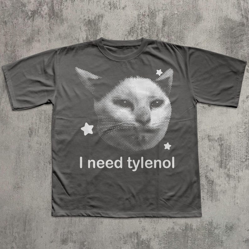 May include: Dark gray t-shirt featuring a white, pixelated cat face with three white stars. The text "I need tylenol" is printed in white below the cat graphic. The shirt is made of a soft material.