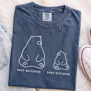 May include: A blue short-sleeved t-shirt featuring a white line-art graphic of two bears. The larger bear is labeled "bear maximum" and the smaller bear is labeled "bear minimum". The shirt has a crew neckline and a Comfort Colors brand label.