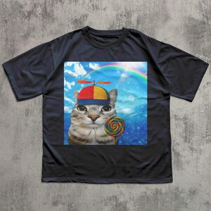 May include: Navy blue t-shirt featuring a whimsical graphic of a cat wearing a propeller hat and holding a lollipop. The cat is set against a backdrop of a rainbow and blue sky, creating a playful and colorful design.