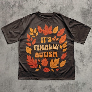 Peut inclure: T-shirt marron foncé avec l'inscription "IT'S FINALLY AUTISM" dans une police rétro, entourée d'une couronne de feuilles d'automne orange et jaune. Le t-shirt a des manches courtes et un col rond.