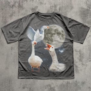Op de afbeelding: Een antracietgrijs T-shirt met korte mouwen en een grafische print van drie witte ganzen tegen een donkere nachtelijke hemel. E&eacute;n gans vliegt met gespreide vleugels, &eacute;&eacute;n staat op de grond en &eacute;&eacute;n gans snatert met open snavel bij de volle maan.
