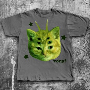 May include: Gray t-shirt featuring a green cat head with multiple eyes and antennae. The cat graphic is surrounded by green stars and the word "vorp?" is printed below the cat.
