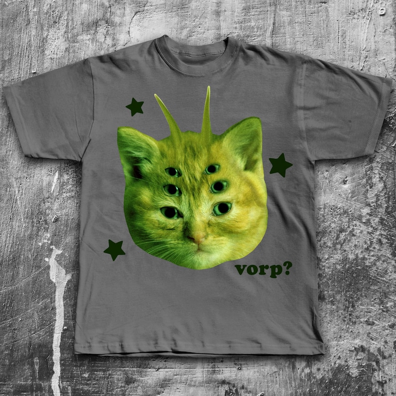 May include: Gray t-shirt featuring a green cat head with multiple eyes and antennae. The cat graphic is surrounded by green stars and the word "vorp?" is printed below the cat.
