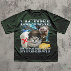 May include: Dark green t-shirt with the words "LACTOSE INTOLERANT" in silver lettering. The graphic features a cat wearing a helmet, a cow, a milk bottle with a red X, and a piece of cheese.