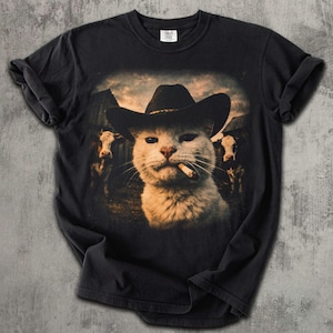 May include: Black t-shirt featuring a humorous graphic of a cat wearing a cowboy hat and smoking a cigarette. The cat is set against a backdrop of a barn and cows, creating a Western theme. The shirt is made of a soft material.