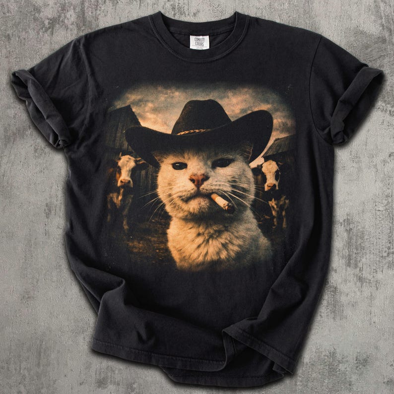 May include: Black t-shirt featuring a humorous graphic of a cat wearing a cowboy hat and smoking a cigarette. The cat is set against a backdrop of a barn and cows, creating a Western theme. The shirt is made of a soft material.