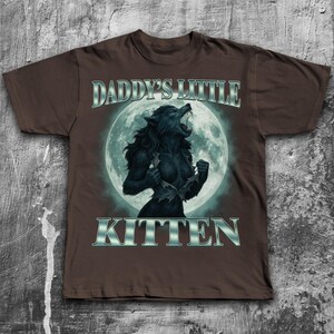 Pode incluir: Camiseta marrom com um gr&aacute;fico de lobisomem em frente a uma lua cheia. O texto "DADDY'S LITTLE KITTEN" &eacute; exibido acima e abaixo do gr&aacute;fico em prata. O lobisomem &eacute; preto e est&aacute; rasgando a camisa.