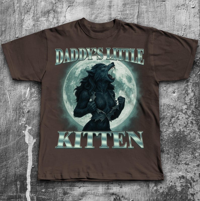 Pode incluir: Camiseta marrom com um gr&aacute;fico de lobisomem em frente a uma lua cheia. O texto "DADDY'S LITTLE KITTEN" &eacute; exibido acima e abaixo do gr&aacute;fico em prata. O lobisomem &eacute; preto e est&aacute; rasgando a camisa.