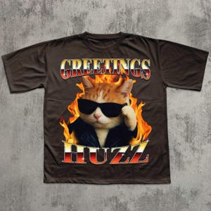 Op de afbeelding: Bruin t-shirt met een afbeelding van een kat met zonnebril en leren jas, omringd door vlammen. De tekst "GREETINGS" en "HUZZ" is in een opvallend lettertype over het ontwerp weergegeven.