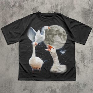 Op de afbeelding: Een zwart t-shirt met korte mouwen en een grafische print van drie witte ganzen tegen een nachtelijke hemel. E&eacute;n gans vliegt met gespreide vleugels, &eacute;&eacute;n staat op de grond en &eacute;&eacute;n gans snatert met de snavel open bij een grote volle maan.