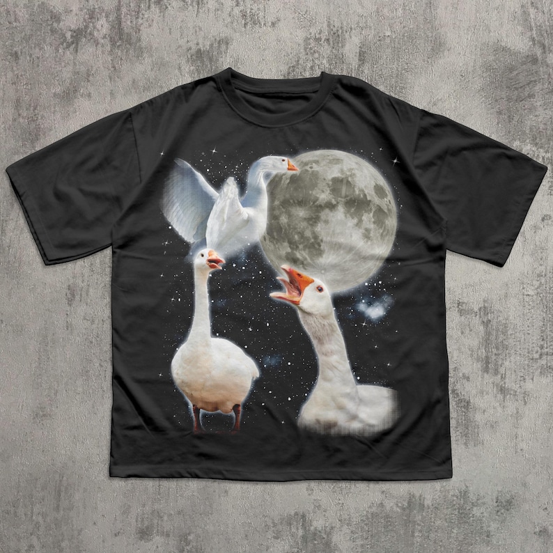 Op de afbeelding: Een zwart t-shirt met korte mouwen en een grafische print van drie witte ganzen tegen een nachtelijke hemel. E&eacute;n gans vliegt met gespreide vleugels, &eacute;&eacute;n staat op de grond en &eacute;&eacute;n gans snatert met de snavel open bij een grote volle maan.