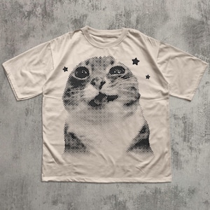 May include: Cream-colored t-shirt featuring a large, halftone image of a cat's face. The cat's expression is curious, with three black star graphics around the cat's head. The shirt has short sleeves and a crew neck.