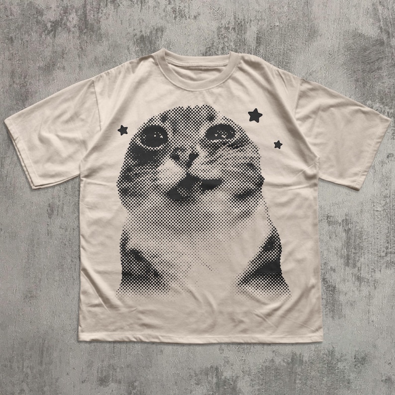 May include: Cream-colored t-shirt featuring a large, halftone image of a cat's face. The cat's expression is curious, with three black star graphics around the cat's head. The shirt has short sleeves and a crew neck.