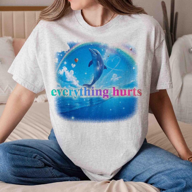 May include: A light gray t-shirt featuring a vibrant graphic of a dolphin leaping from the ocean, set against a blue sky with clouds and a rainbow. The text "everything hurts" is displayed in colorful letters.