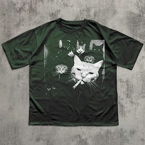 May include: Dark green t-shirt featuring a black and white graphic of several cat faces, with one cat smoking a cigarette. The design includes various cat faces in different sizes and expressions, creating a unique and artistic look.