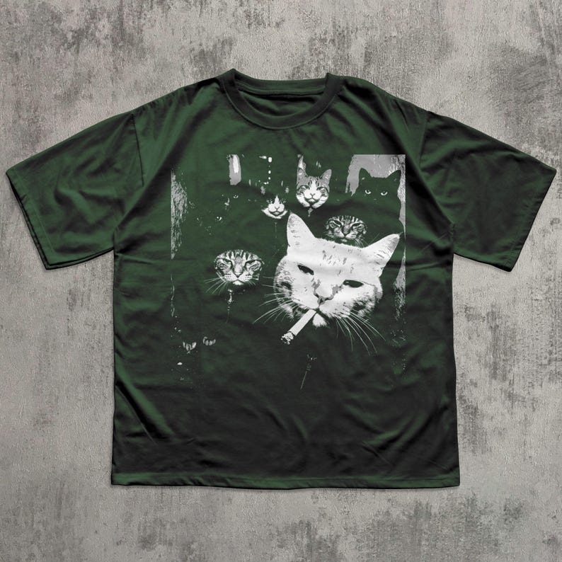 May include: Dark green t-shirt featuring a black and white graphic of several cat faces, with one cat smoking a cigarette. The design includes various cat faces in different sizes and expressions, creating a unique and artistic look.