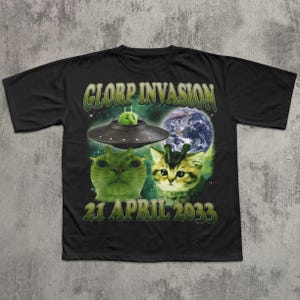以下が含まれることがあります： 黒いTシャツ。デザインには「GLORP INVASION」の文字、UFO、猫のような宇宙人2体、日付「21 APRIL 2033」が含まれています。デザインは緑と青の色合いです。