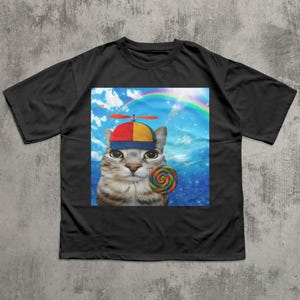 May include: Black t-shirt featuring a whimsical graphic of a cat wearing a propeller hat and holding a colorful lollipop. The background includes a rainbow and a blue sky with clouds. The cat's expression is curious.