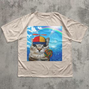 May include: Beige t-shirt featuring a whimsical graphic of a cat wearing a propeller hat and holding a colorful lollipop. The cat is set against a backdrop of a rainbow and blue sky, creating a playful and unique design.