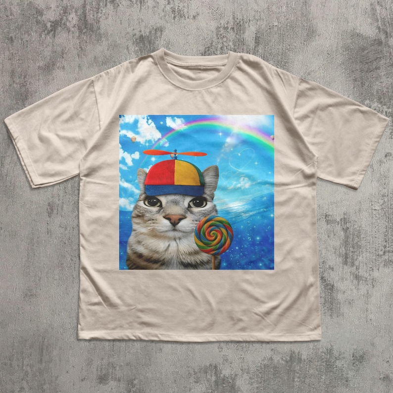 May include: Beige t-shirt featuring a whimsical graphic of a cat wearing a propeller hat and holding a colorful lollipop. The cat is set against a backdrop of a rainbow and blue sky, creating a playful and unique design.