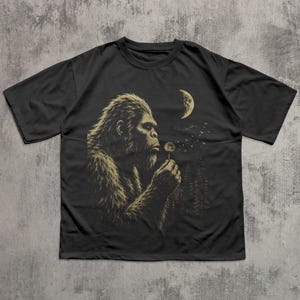 May include: A black short-sleeve t-shirt featuring a graphic print of a hairy creature resembling a Bigfoot holding a dandelion. The design includes a crescent moon and a forest silhouette in the background, rendered in muted gold tones.