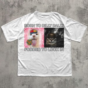May include: White t-shirt with the text "BORN TO DILLY DALLY FORCED TO LOCK IN". The shirt features two cat images: one with a lollipop and the other wearing a helmet.