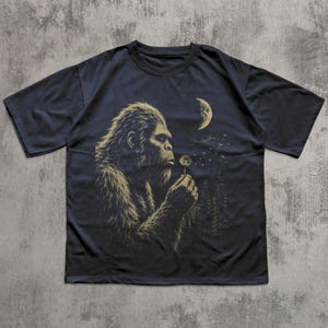 May include: A dark navy blue short-sleeve t-shirt featuring a graphic print of a hairy creature resembling Bigfoot blowing on a dandelion. The design includes a crescent moon and a forest silhouette in the background in muted gold tones.