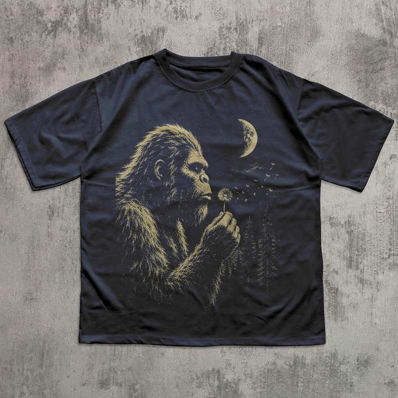 May include: A dark navy blue short-sleeve t-shirt featuring a graphic print of a hairy creature resembling Bigfoot blowing on a dandelion. The design includes a crescent moon and a forest silhouette in the background in muted gold tones.