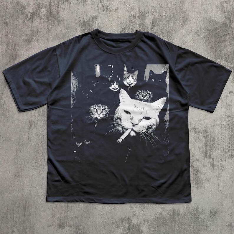 May include: Dark navy blue t-shirt featuring a black and white graphic of several cat faces, with one cat smoking a cigarette. The design has a vintage, slightly distressed look, with a grunge aesthetic.