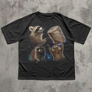 May include: Black t-shirt featuring a whimsical graphic of three raccoons. One raccoon is holding a trash can, while the others look up. The design is set against a cosmic background with stars and nebulas.