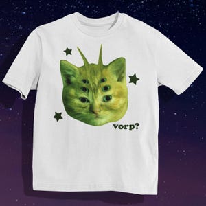 May include: White t-shirt featuring a green cat-like creature with multiple eyes and spiky antennae. The design includes dark green star shapes and the word "vorp?" in black. The shirt has short sleeves and a crew neck.