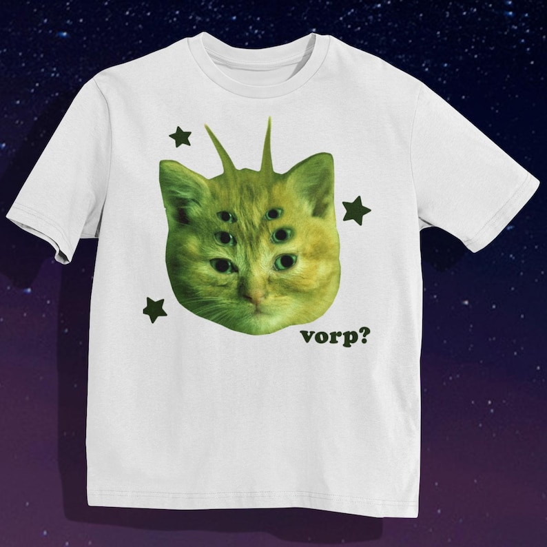 May include: White t-shirt featuring a green cat-like creature with multiple eyes and spiky antennae. The design includes dark green star shapes and the word "vorp?" in black. The shirt has short sleeves and a crew neck.