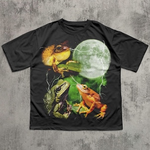 Può includere: T-shirt nero con una grafica di tre rane, una luna piena e fulmini. Le rane sono verdi e arancioni, con un bagliore verde intorno alla luna. Il disegno è stampato sulla parte anteriore della maglietta.