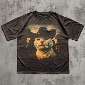 May include: Dark brown t-shirt featuring a graphic of a cat wearing a cowboy hat and smoking a cigarette. The cat is in front of a mountain scene with cows. The shirt has short sleeves and a crew neck.