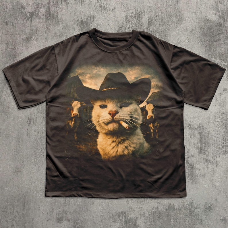 May include: Dark brown t-shirt featuring a graphic of a cat wearing a cowboy hat and smoking a cigarette. The cat is in front of a mountain scene with cows. The shirt has short sleeves and a crew neck.