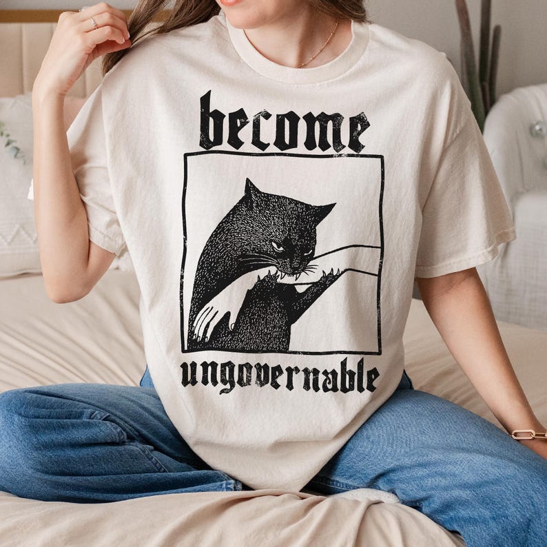 Op de afbeelding: Cr&egrave;mekleurig T-shirt met een zwarte kattenafbeelding in een zwart rechthoekig kader. De tekst "become" staat boven de kat en "ungovernable" eronder. De afbeelding toont een grote en een kleine kat met een hand.