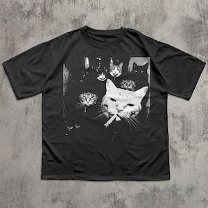 May include: Black t-shirt featuring a graphic of several cats, with one large white cat smoking a cigarette. The other cats are in the background, with various expressions. The design is in black and white.