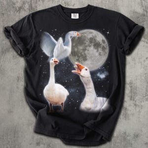 Op de afbeelding: Een zwart T-shirt met korte mouwen en een grafische print van drie witte ganzen tegen een sterrenhemel met een grote volle maan. E&eacute;n gans vliegt, &eacute;&eacute;n staat en &eacute;&eacute;n heeft de snavel open alsof hij luidkeels aan het gakkern is.