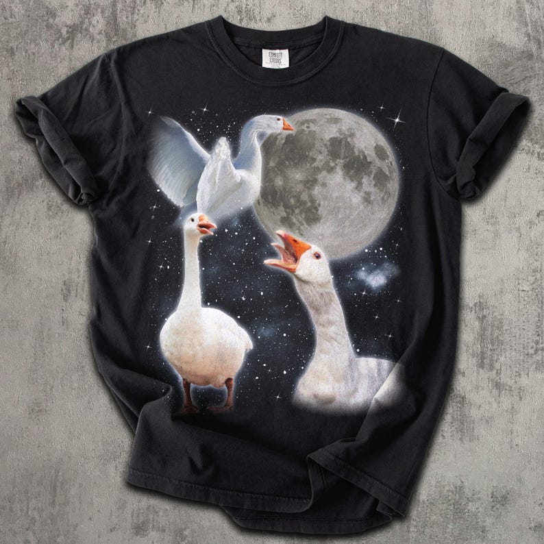 Op de afbeelding: Een zwart T-shirt met korte mouwen en een grafische print van drie witte ganzen tegen een sterrenhemel met een grote volle maan. E&eacute;n gans vliegt, &eacute;&eacute;n staat en &eacute;&eacute;n heeft de snavel open alsof hij luidkeels aan het gakkern is.