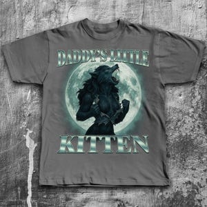 Pode incluir: Camiseta cinza com um gr&aacute;fico de lobisomem em frente a uma lua cheia. O texto "DADDY'S LITTLE KITTEN" &eacute; impresso acima e abaixo do gr&aacute;fico em uma fonte met&aacute;lica prateada. O lobisomem est&aacute; rasgando sua camisa.