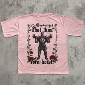 May include: A light pink t-shirt featuring a medieval knight lifting dumbbells. The knight is surrounded by a floral design with the text "Good sir dost thou even hoist?" in a gothic font. The shirt is made of a soft material.