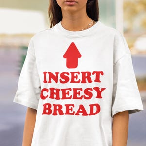 May include: White t-shirt with the text "INSERT CHEESY BREAD" in red, along with a red arrow pointing upwards. The shirt has short sleeves and a casual fit.