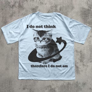 May include: Light blue t-shirt featuring a black and white illustration of a kitten inside a curved line with a star. The text "I do not think" is above the kitten, and "therefore I do not am" is below.