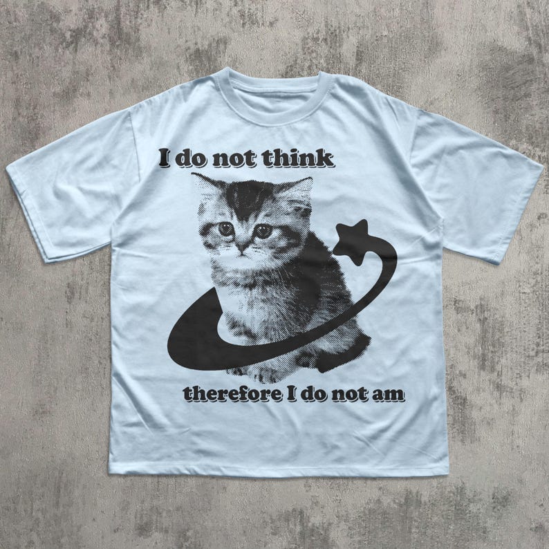 May include: Light blue t-shirt featuring a black and white illustration of a kitten inside a curved line with a star. The text "I do not think" is above the kitten, and "therefore I do not am" is below.