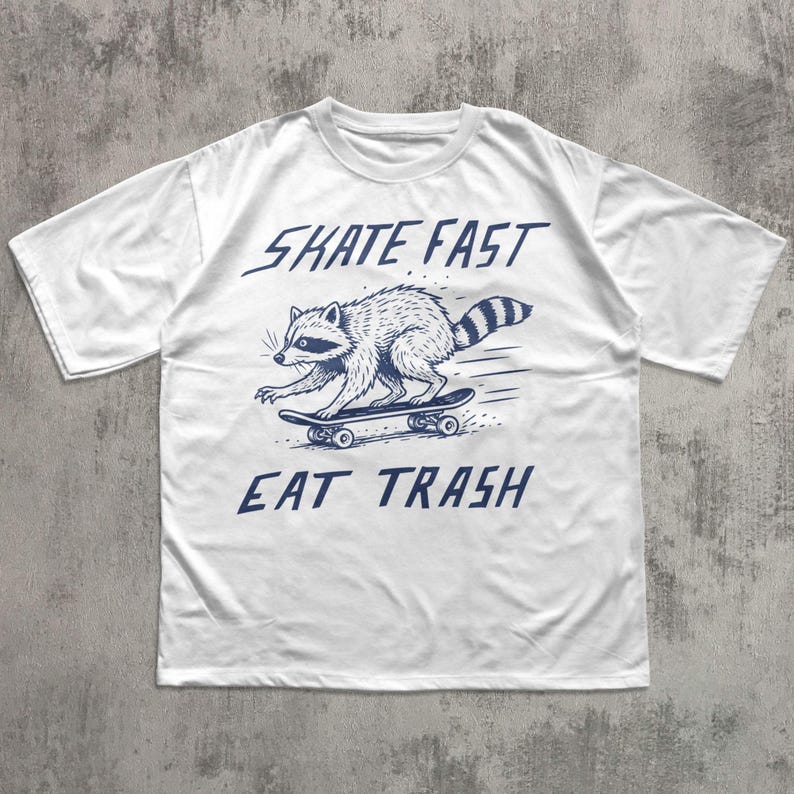 May include: White t-shirt featuring a raccoon skateboarding. The raccoon is illustrated in blue, with the words "SKATE FAST EAT TRASH" above and below the image. The shirt has short sleeves and a crew neck.