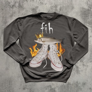 May include: Dark grey sweatshirt featuring a fish wearing a gold crown, standing on white high-top trainers. The word "fih" is above the fish in white, with flames around the trainers.